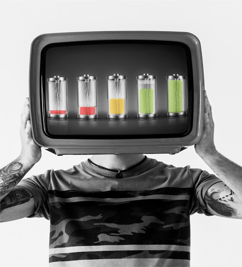 A person in a camouflage t-shirt holds an analog television displaying a row of light batteries in place of their head, symbolizing a concept of ideas, energy, or technology-driven thought.