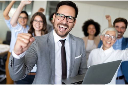 A group of jubilant colleagues celebrate a success in an office setting, with a man in a suit at the forefront, looking directly at the camera, fist raised victoriously, evoking a sense of shared achievement and team spirit.